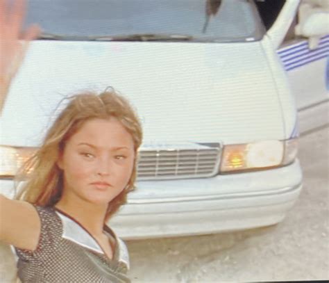 a woman standing in front of a white car with her hand up to the camera