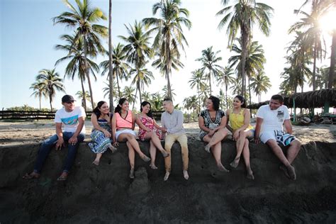 Group Of Smiling Friends Sits On A Stone Wall At The Sunny Beach Near
