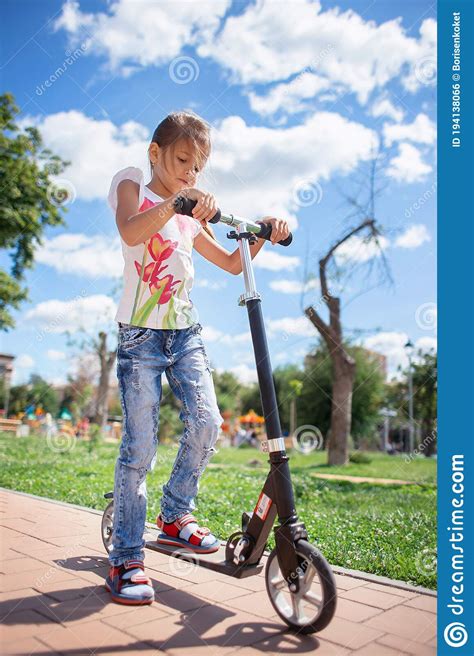 A Girl In Torn Fashionable Jeans Is Riding A Scooter In A Summer Park