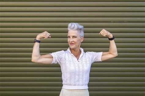 Senior Woman Flexing Muscles While Standing Against Shutter — Grey Hair