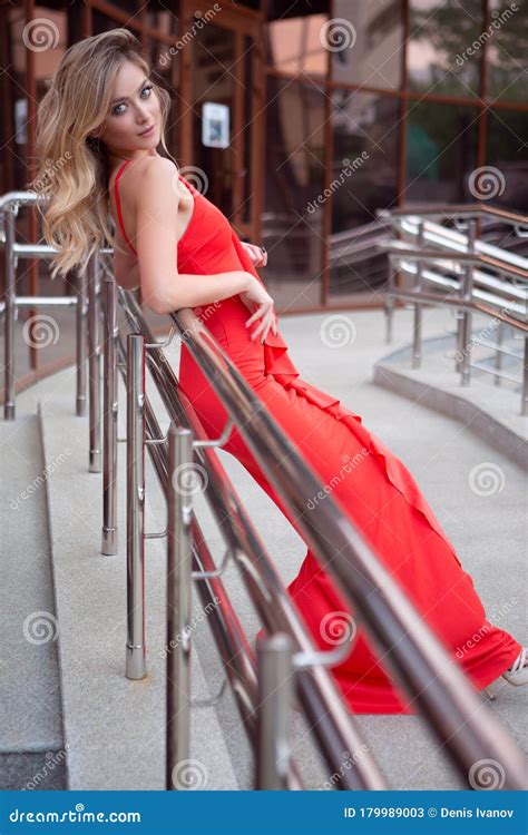 an elegant sexy woman in a tight red dress poses leaning against the railing of a street