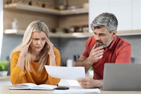 Stressed Husband And Wife Sitting At Kitchen Table Reading Documents