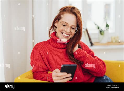 Happy Beautiful Red Haired Girl In Glasses And Red Sweater Looking At Smartphone In Her Hand