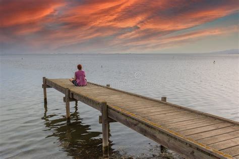 Red Haired Woman Sitting On A Wooden Dock Watching The Sunset Stock