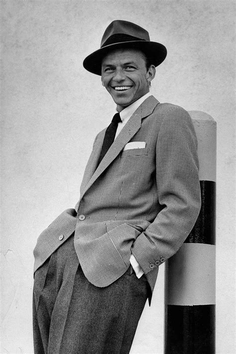 A Black And White Photo Of A Man In A Suit Leaning On A Pole Smiling