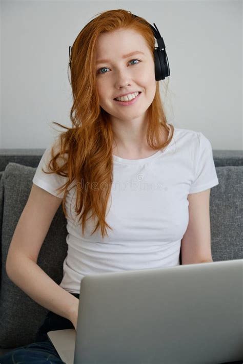 a cute redhead girl sits on a sofa in a white t shirt and headphones with a laptop and smiles
