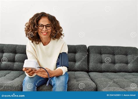Mature Hispanic Woman Using Blood Pressure Monitor Sitting On The Sofa