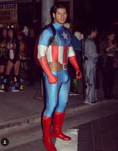 A Man Dressed As Captain America Stands On The Street At Night With People In Costume