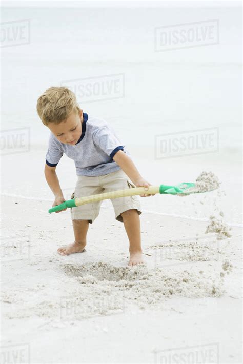 Young Boy Standing On Beach Digging In Sand With Shovel Looking Down