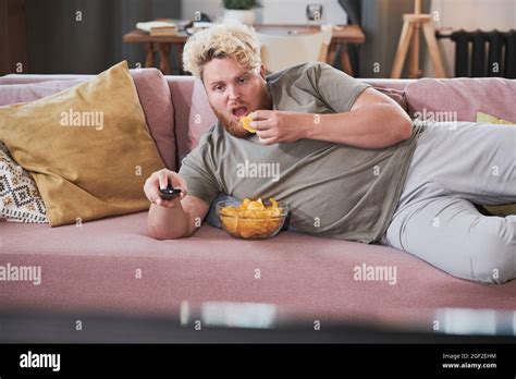Lazy Overweight Man Lying On The Sofa Eating Chips And Watching TV At