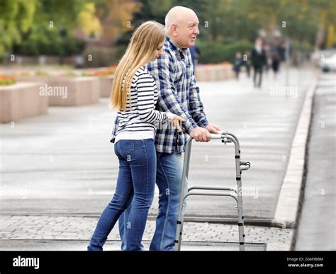Young Woman Helping Elderly Man With Walking Frame To Cross The Road