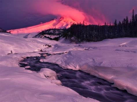 A Mountain Covered In Snow Next To A River Under A Pink Sky With Clouds