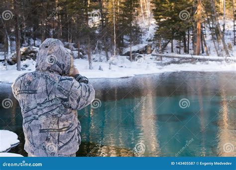 A Man In Camouflage Winter Clothes Examines The Lake With A Colorful