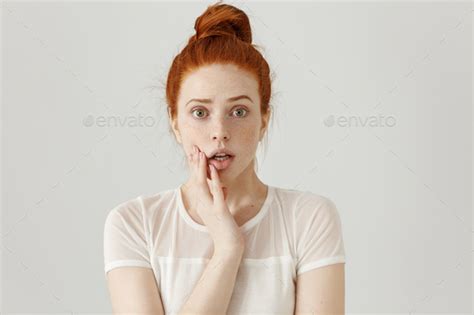 Frightened Startled Young Redhead Freckled Female Raising Eyebrows Keeping Hand On Her Cheek