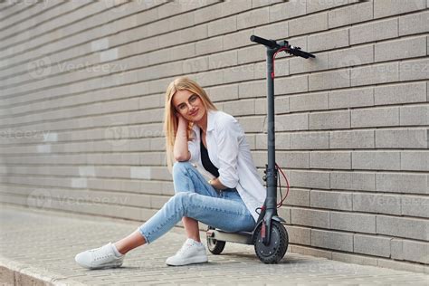 Beautiful Blonde In Casual Clothes Sitting On Electric Schooter Outdoors At Sunny Daytime