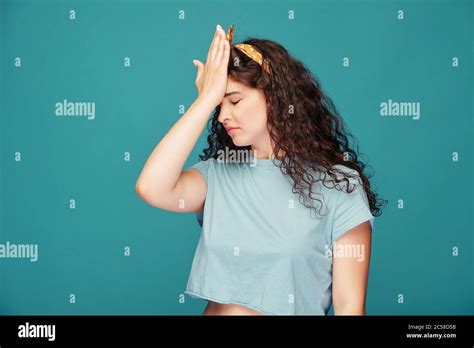 upset curly haired girl making facepalm gesture displaying disappointment against blue