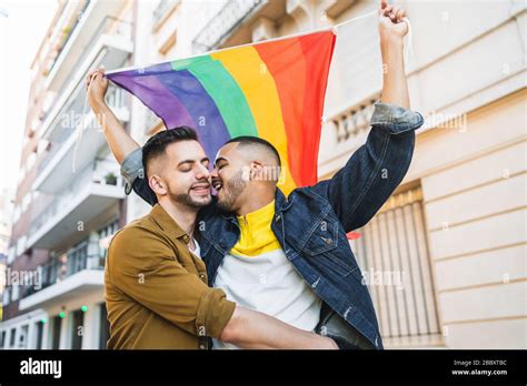 Portrait Of Young Gay Couple Embracing And Showing Their Love With Rainbow Flag At The Street