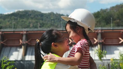 engineer mother wearing helmet to her daughter against background of dam with hydroelectric