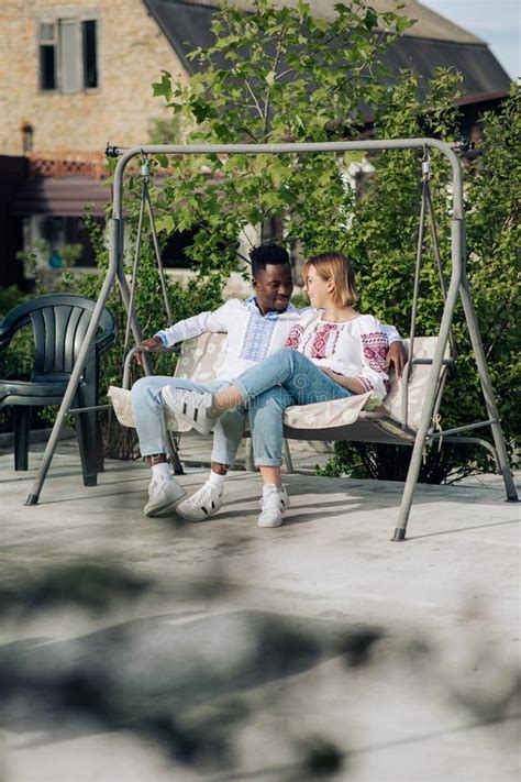 Interracial Couple Sits On Bench Swing In Garden Dressed In Ukrainian Embroidered Shirts Stock