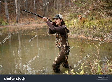Woman Duck Hunter Wearing Camo Waders With Rifle In Pond And Dog In
