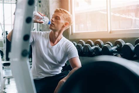 Young Caucasian Man At A Gym Sit On Bench And Drinking Water From