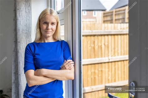 Blond Mature Woman Leaning With Arms Crossed On Glass Door At Home