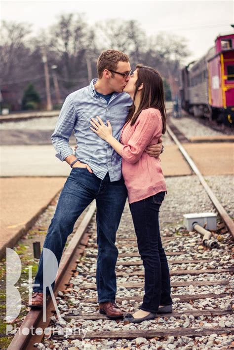 A Man And Woman Standing On Train Tracks With Their Arms Around Each Other As They Kiss