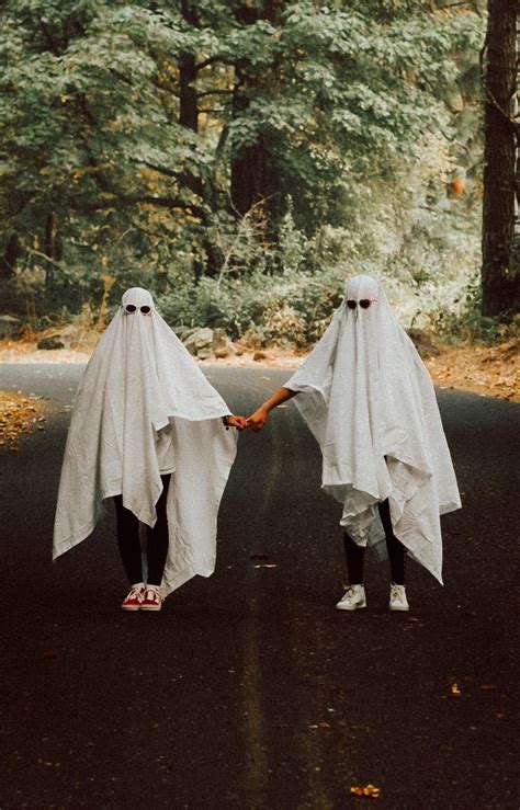 Two People In White Ghost Costumes Holding Hands On A Road With Trees