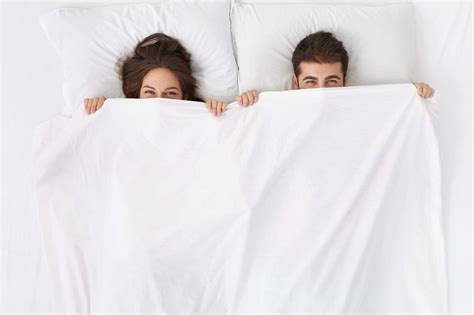Funny married couple lying in bed and hiding under white blanket, looking at camera with eyes ...