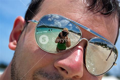 Close Up Of A Man Wearing Reflective Sunglasses In A Tropical Beach With Reflection Of The Woman