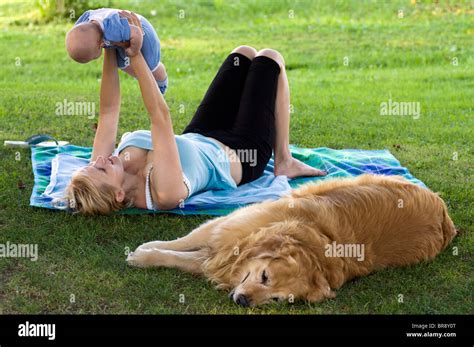Mother Playing With Her Baby In The Park With Golden Retriever Lying