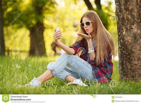 Young Beautiful Woman Takes Selfie On Cell Phone Sitting On Grass In Summer City Park Blow Kiss