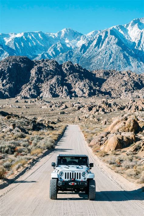 A White Jeep Driving Down A Dirt Road In Front Of Snow Capped