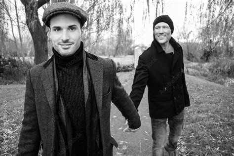 Black And White Portrait Of Male Gay Couple Holding Hands And Smiling As They Walk Through Park