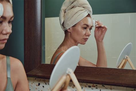 Young Woman Using Jade Facial Roller For Face Massage Sitting In Bathroom Looking In The Mirror