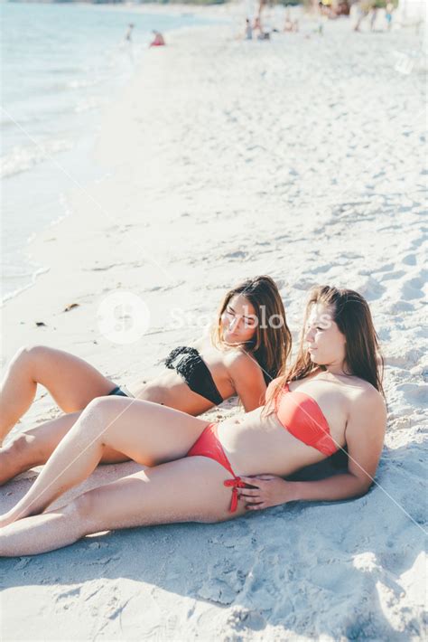 Two Young Women Millennials Lying Down At The Beach Tanning Relaxing Bonding Interaction