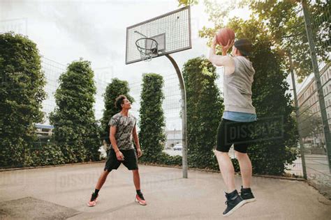 Young Man Taking Jump Shot With Friend On Basketball Court Streetball