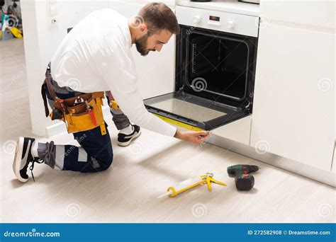 Repairman Examining Oven With Screwdriver In Kitchen With Tool Case