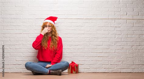 Young Redhead Woman Sitting Over Brick Wall Wearing Christmas Hat Smelling Something Stinky And