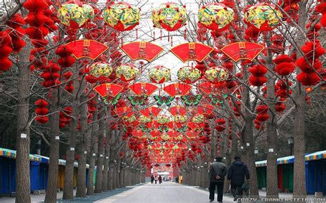 Two People Walking Down A Street Lined With Trees And Red Lanterns