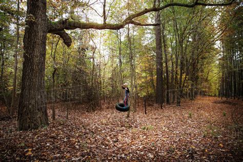 Boy Standing On Tire Swing Hanging From Tree In Forest Stock Photo