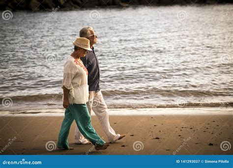 Senior Adult Couple Walk Together On The Shore With Ocean In Background Enjoy The Leisure