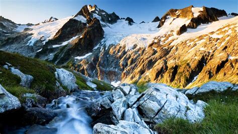 Rock Greenery Slope Mountains With Snow Sunlight In Blue Sky Background