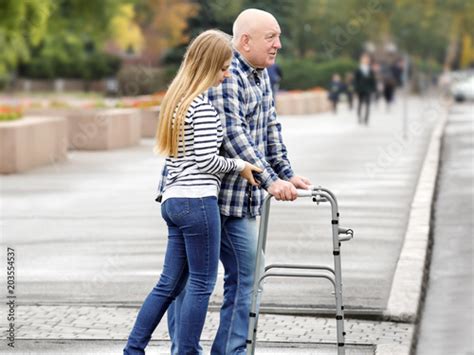 Young Woman Helping Elderly Man With Walking Frame To Cross The Road