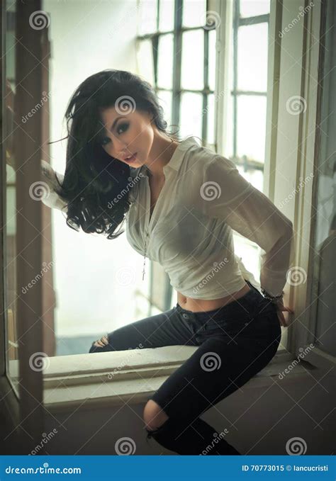 Attractive Brunette In White Tight Fit Shirt And Black Ripped Jeans Posing Sitting On Window