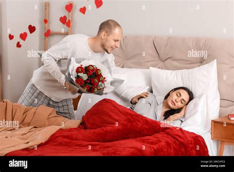 Young Man Greeting His Sleeping Wife With Flowers In Bedroom On