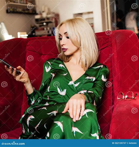 Beautiful Blond Woman In Green Dress Sitting On Red Sofa And Typing Something On Her Phone Stock