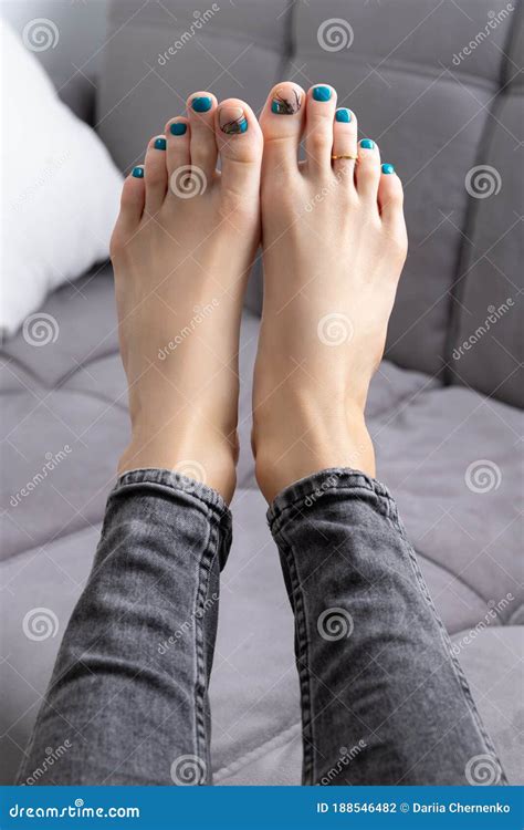 Womans Feet In Jeans With Pedicure On Gray Sofa At Home Stock Photo