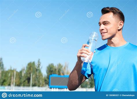 Young Sporty Man Drinking Water From Bottle At Stadium On Sunny Day