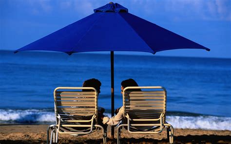 Couple Sitting On White Steel Folding Bed Near In The Beach Under Blue
